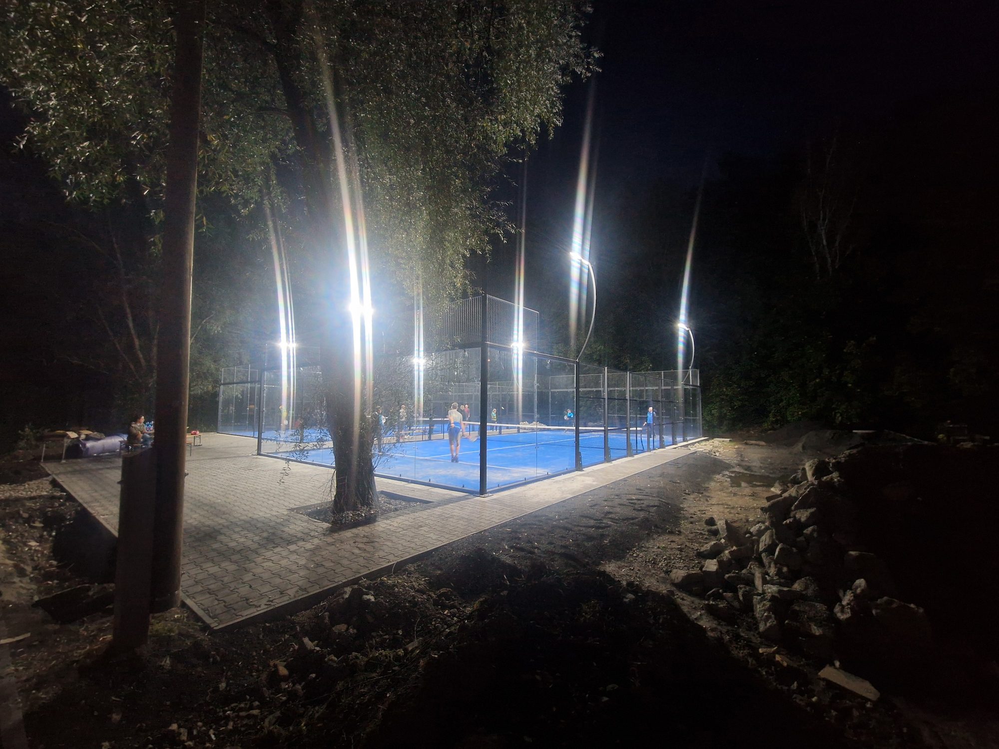 Hráči na osvětleném padelovém kurtu za tmy, pohled od dlážděného chodníku