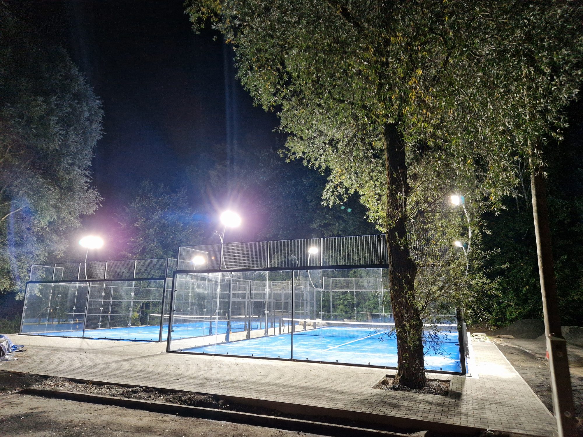 Osvětlené prosklené padelové kurty s modrým povrchem, v popředí strom
